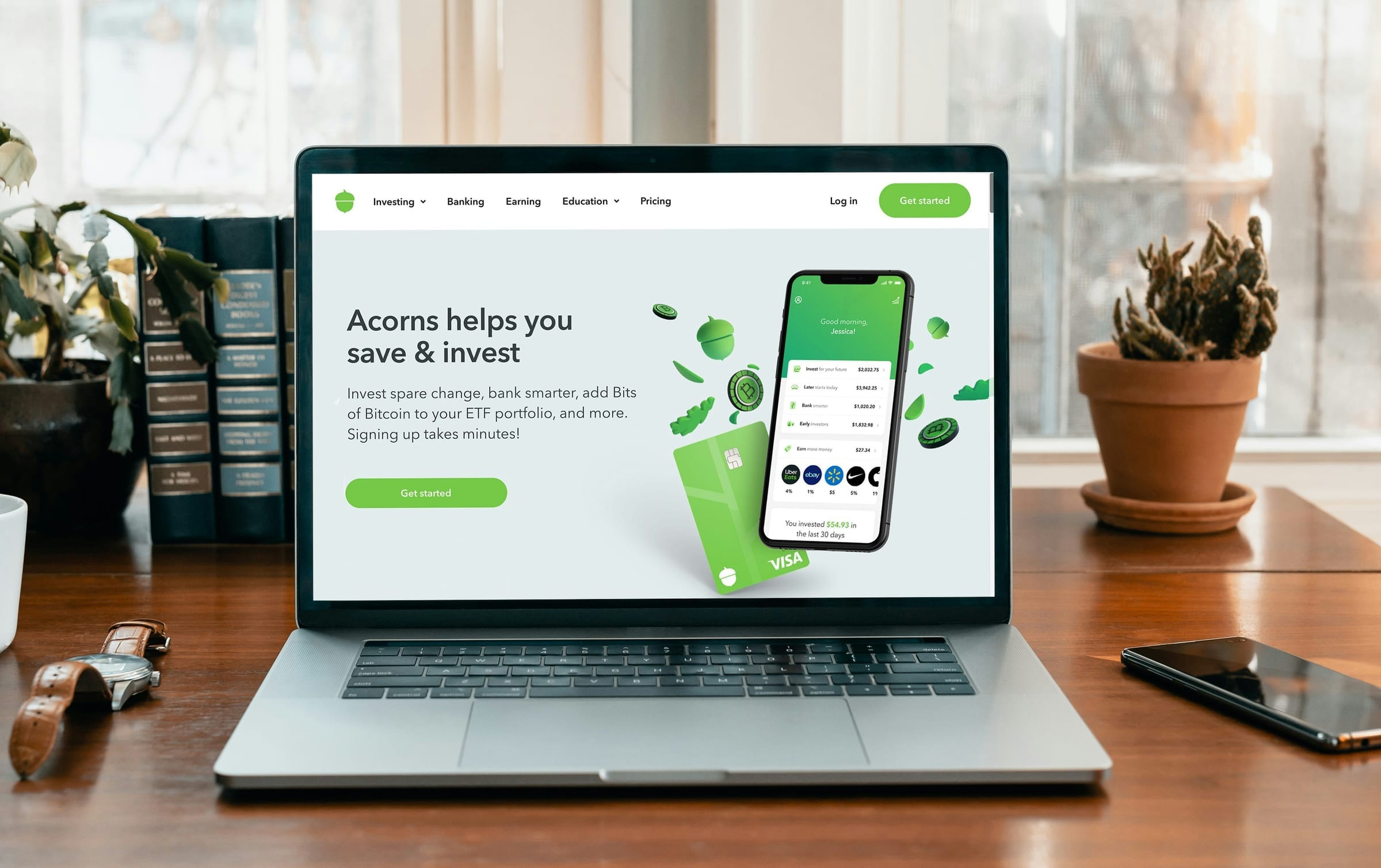 An open laptop on a wooden desk showing the landing page of a personal finance app, with a phone, a watch, books and plants around it