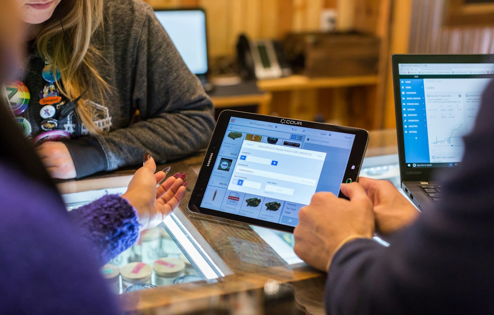 Two people at a store counter: one adds a product on a tablet while the laptop beside shows a sales dashboard